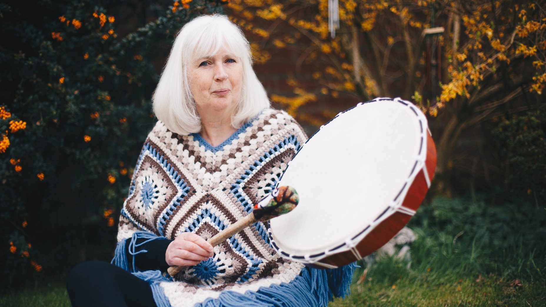 White headed woman smiling and beating a white and red shamanic drum in nature