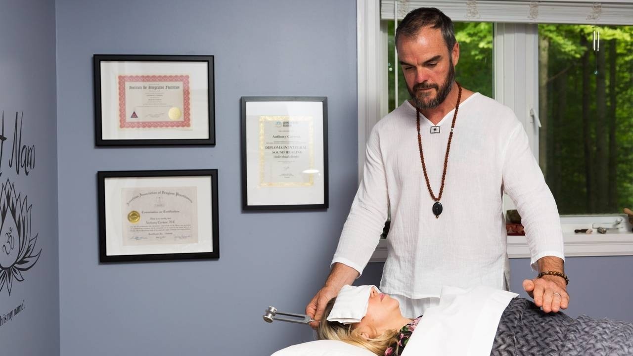 Sound healing practitioner man, holding a weighted tuning for on his client's head, and the other hand hovering above the solar plexus area