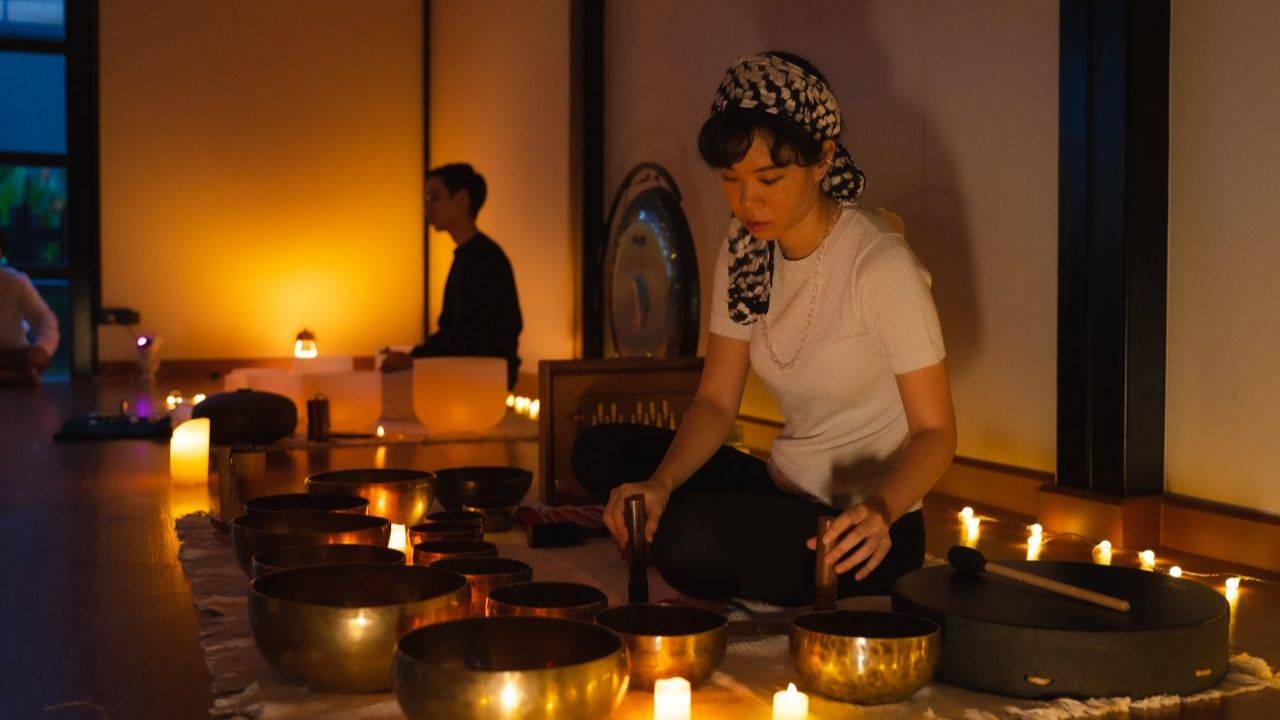 Man holding a tibetan singing bowl and sitting in the lotus position, surrounded by many other crystal and tibetan singing bowls