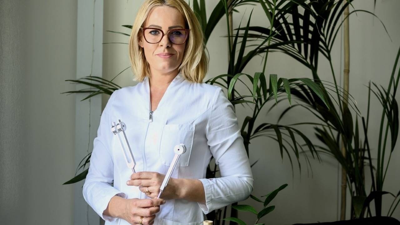 Blonde woman with glasses holding tuning forks next to a large indoor plant