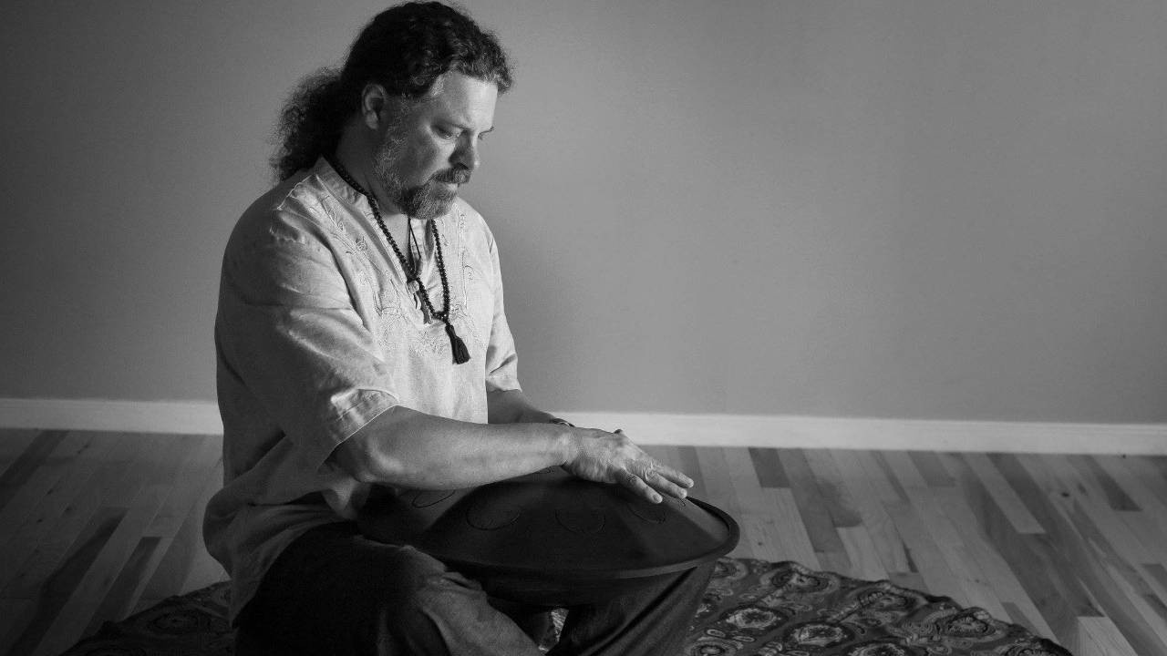 Black and white image of a man sitting down and playing a hand drum