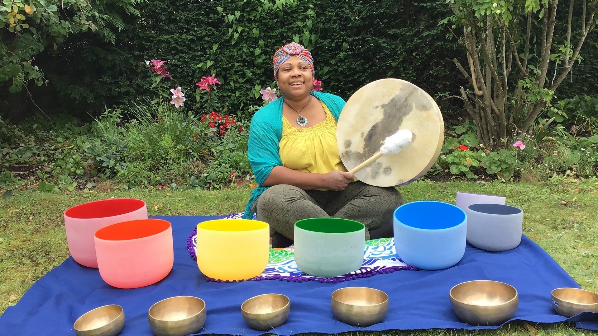 Woman beating a hand held drum and sitting in front of colourful crystal bowls in nature