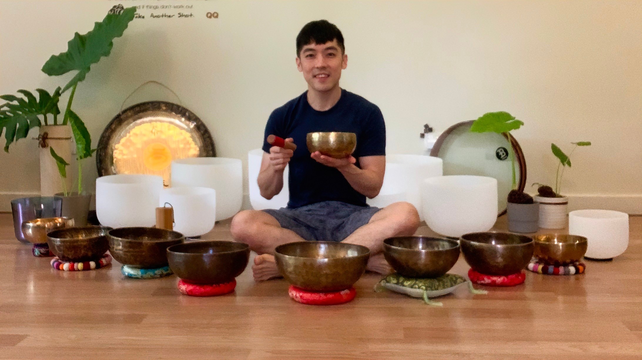 Man holding a tibetan singing bowl and sitting in the lotus position, surrounded by many other crystal and tibetan singing bowls