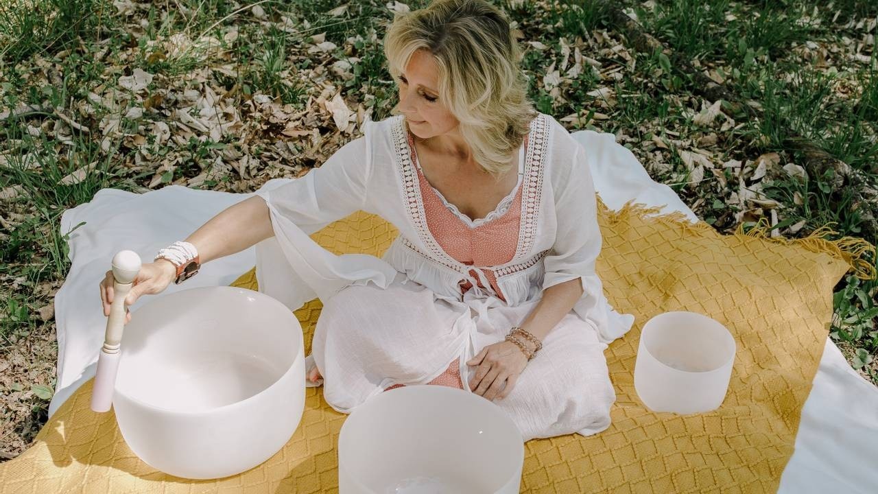 Woman playing the rim of a large white crystal singing bowl outdoors in nature on yellow and white blankets