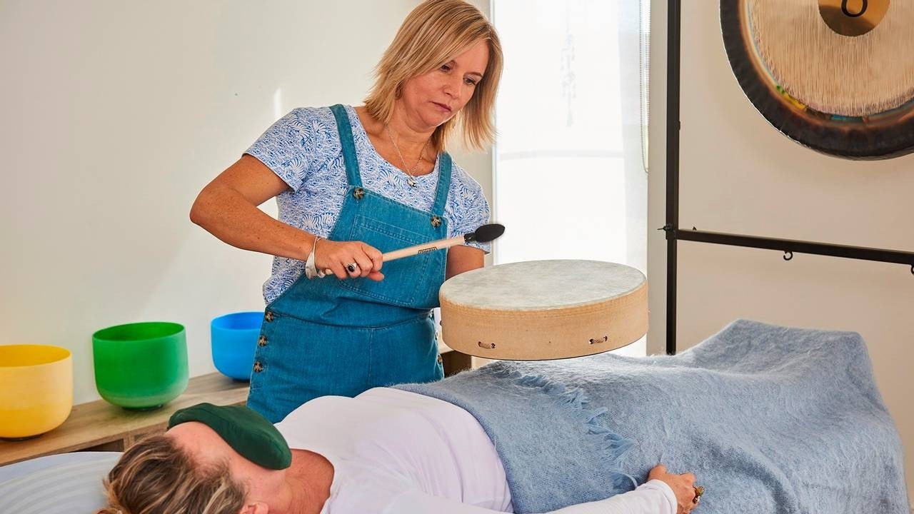 Blonde woman sound healing practitioner, beating a drum above her client's sacral area