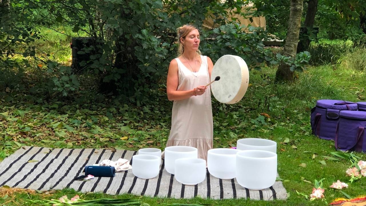 Woman beating a drum in nature next to crystal singing bowls