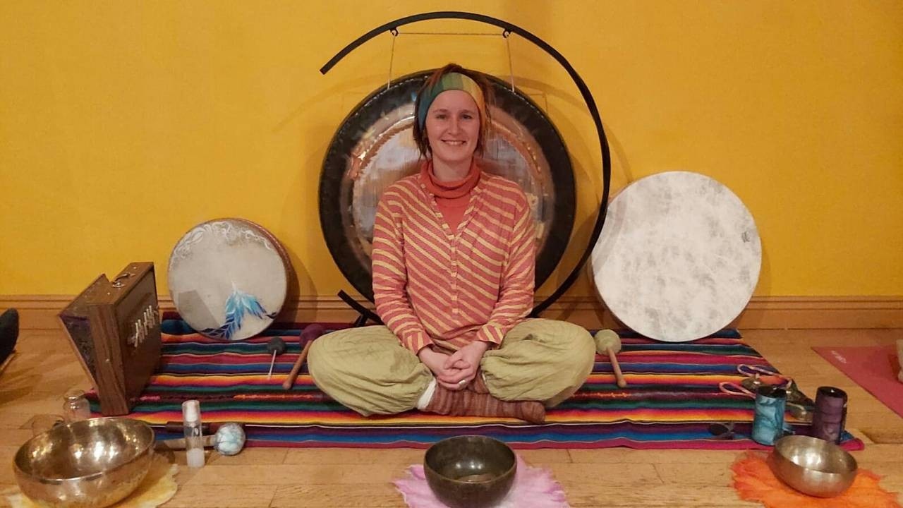 Woman smiling and sitting in the lotus position, surrounded by sound healing instruments indoors