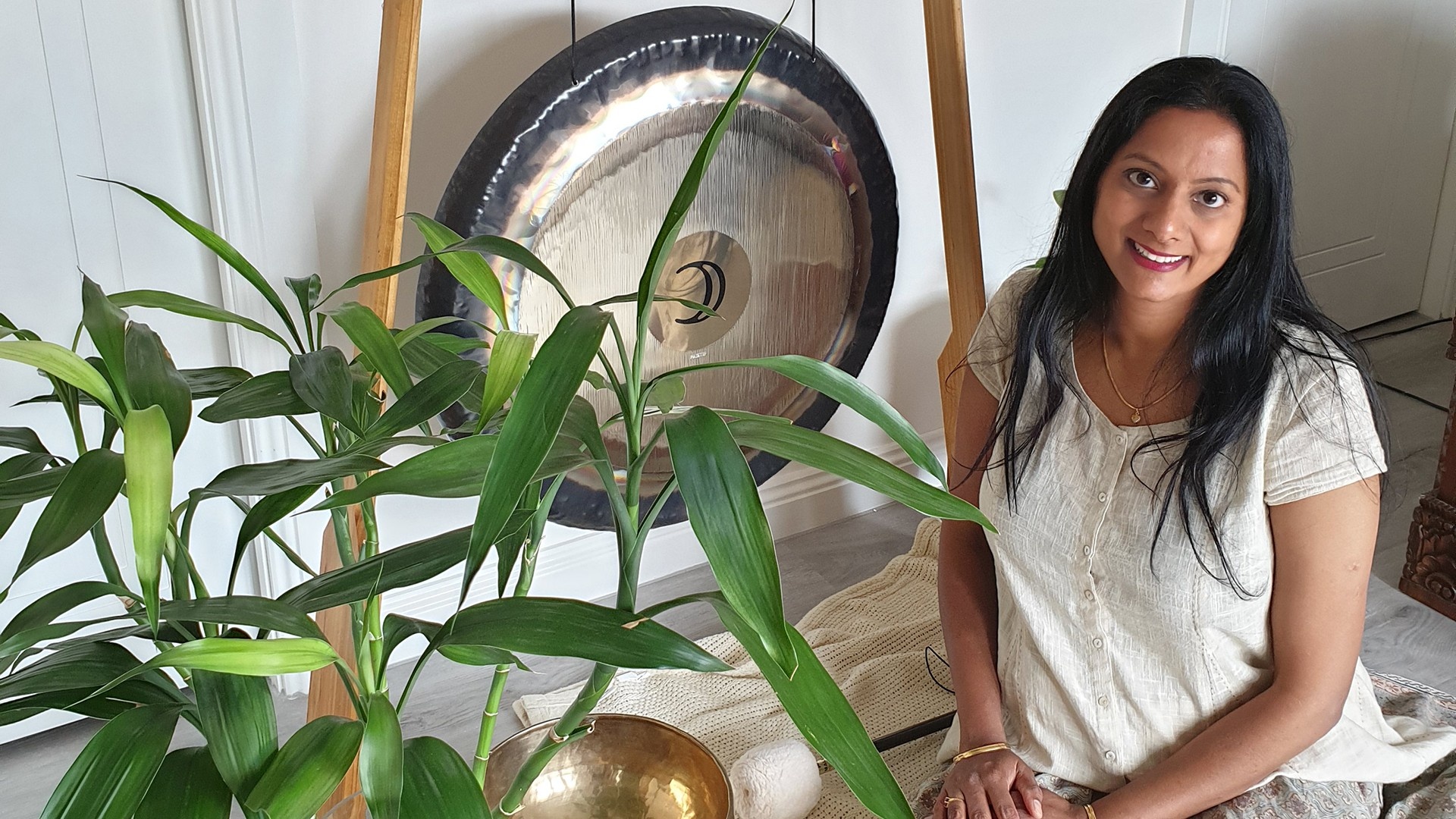 Sound healing practitioner sitting next to a big standing gong, a large plant, and a tibetan singing bowl